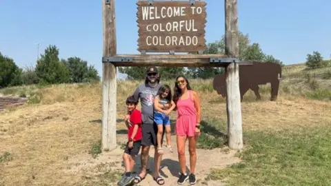 Ciara Fernandez Faber Colorado mother Ciara Fernandez Faber, wearing a short pink romper, stands with her husband, son and daughter under a sign that reads 'Welcome to Colourful Colorado' - with grass, shrubbery and a bison art piece in the background