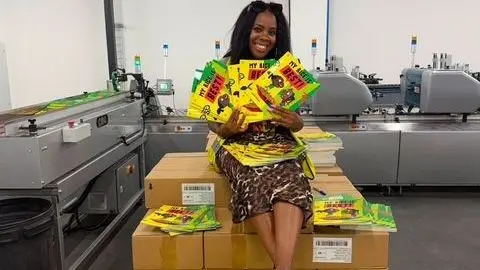 Selina Brown Children's author Selina Brown, wearing a black and brown animal pattern dress, holds five copies of her book, My Rice Is Best, while sitting atop a pile of boxes holding more copies