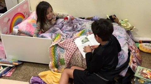 Ciara Fernandez Faber A young boy clad in black shorts and a black top sits on the ground, holding a book on the edge of his sister's bed as he reads it to her; she sits under a purple blanket, a rainbow visible on the headboard