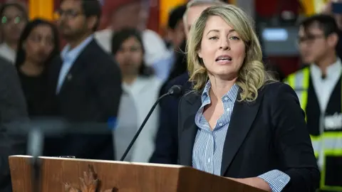 Getty Images An image showing Melanie Joly speaking at a prodium. She is wearing a black suit jacket and a striped button-up blue collared shirt. She has shoulder length, wavy blonde hair.