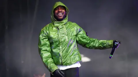 Getty Images Skepta performs on stage wearing a green jacket, black gloves and black trousers.  