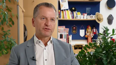 ORF Gerald Tatzgern, a man with short, grey hair, wearing a light grey suit and a white shirt, sitting in an office with books, pot plants and police memorabilia behind him.