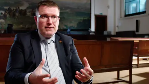 VRT Vincent Guerra, a man with short brown hair and metal-rimmed glasses, wearing a navy suit and a pale shirt and tie, gestures with both hands towards the camera as he is interviewed in a large room, with rows of benches behind him and a large painting on the far wall.