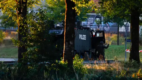 EPA A armed police officer in black heavy duty uniform gestures with his hand as he stands outside an armoured vehicle parked sideways on a road. The photo is taken from a distance, with a patch of grass and trees between the photographer's location and the vehicle