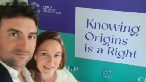 Courtesy of Audrey and Arthur Kermalvezen Audrey and Arthur stand close together in front of a large campaign banner which displays the message “Knowing Origins is a Right” and includes logos associated with child identity advocacy.
