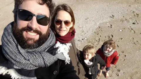 Courtesy of Audrey Kermalvezen A close-up selfie shows four people on a sandy beach, with two adults in the foreground wearing sunglasses, coats, and scarves, and two young children standing below them looking toward the camera, one wearing a red coat, while the other wears a black coat. In the background the sand is covered in shells and footprints. 