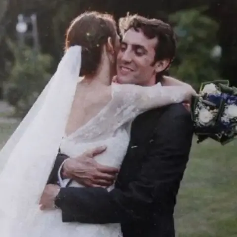 Courtesy of Audrey Kermalvezen A married couple embrace in a grassy area like a garden. The bride, her back to the camera and wearing a white wedding dress with a long white veil, is being lifted and embraced by the groom. He is wearing a dark suit. She is holding bouquet of white and blue flowers. 