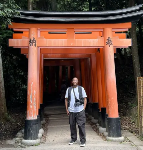 Aden Ibrahim Aden Ibrahim, a man in white T-shirt and black trousers, stands under an orange torii gate in Japan