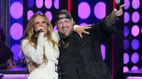 Reuters Jelly Roll in black denim jacket and backwards black baseball cap, with one arm around Carly Pearce and the other holding a microphone and pointing in the air. Carly Pearce is weating a white lace dress and singing into her handheld microphone.