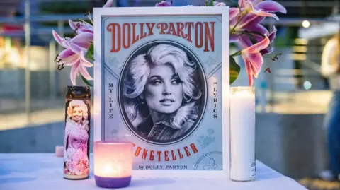 Getty Images An illustration of Dolly Parton with her name stands on a table in front of a vase of pink lilies and behind three candles during the during a Seattle Community Vigil for Dolly Parton on Friday