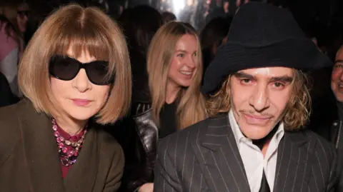 Getty Images Anna Wintour in dark glasses and John Galliano in grey suit and black hat, seated together at a fashion show and looking expressionless at the camera 