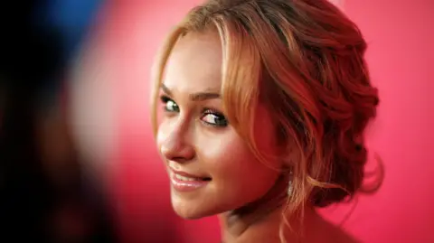 Reuters Hayden Panettiere's head seen turned to look behind her and smile softly, in front of a red background