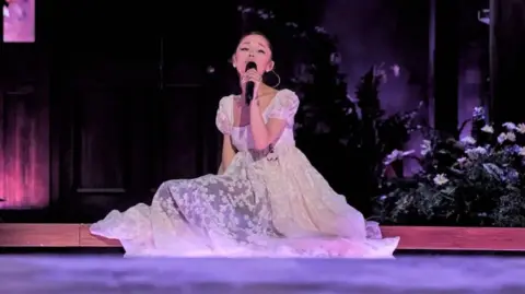 Katia Temkin Ariana Grande sits on the stage, surrounded by flowers, as she sings on her Eternal Sunshine tour