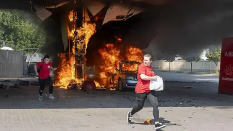 Getty Images Two women in red tops run as flames rise from a car and fuel dispenser following a drone attack by Russia on a petrol station near the city centre of Kharkiv on 3 July