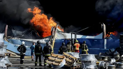 Reuters Wide shot showing Ukrainian firefighters in black and yellow uniforms observing flames rising from the debris of what appears to be a warehouse following a drone strike.