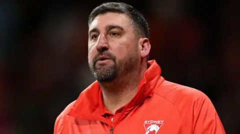 Getty Images A man wears a red jacket emblazoned with a triangular "Sydney Swans" logo.