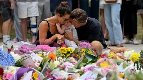 Getty Images A woman with dark hair wearing a black strappy top and a man with dark blonde hair wearing a black shirt hug a young child as they kneel at a floral tribute to the Bondi Beach shooting victims at the Bondi Pavillion in Sydney on 15 December, 2025.
