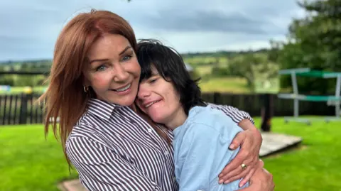 BBC A mother and her son sit in a bright garden. The woman is smiling down fondly at her son as he holds her arm. She has long red hair that is being blown by the wind and is wearing a shirt with stripes. The young man alongside her has a big smile. 