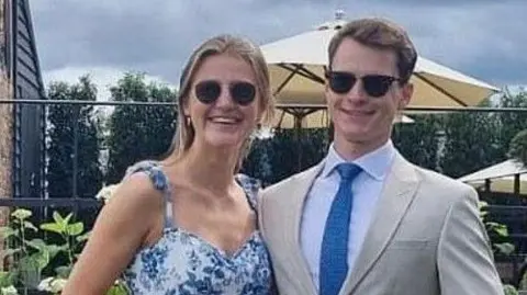 Social Media Alexander Cromie and Marie Ebert stand arm in arm and smile. Alexander is wearing a tan suit and sunglasses. Marie is also wearing sunglasses and a floral dress. 