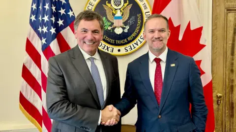 @DLeBlancNB on X An image showing US trade representative Jamieson Greer (right) shaking hands with Canada-US trade minister Dominic LeBlanc to his left. Behind them are a US and Canada flag with the seal for the US trade representative in the middle. Both men are wearing suits and smiling at the camera.