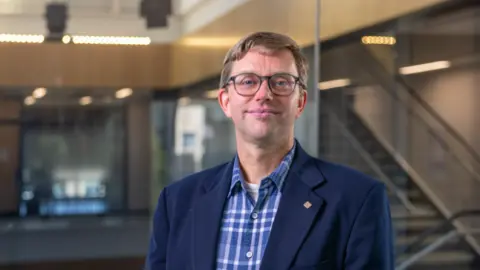 Supplied by the University of Western Ontario An image showing Prof Guikema. He has short dirty blonde hair, and round glasses with thin black frames. He is slightly smiling. He is wearing a navy blue smart coat with a checkered blue button-up shirt underneath. Behind him is a backdrop of an academic building, showing glass panels and stairs that are blurred. 