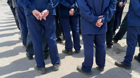 BBC The backs of a line of men wearing a navy uniform. They are standing with one hand holding the other behind their backs. 