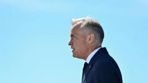 Reuters Mark Carney stands in profile, facing to the left, against a clear light blue sky. He wears a dark blue suit and tie.