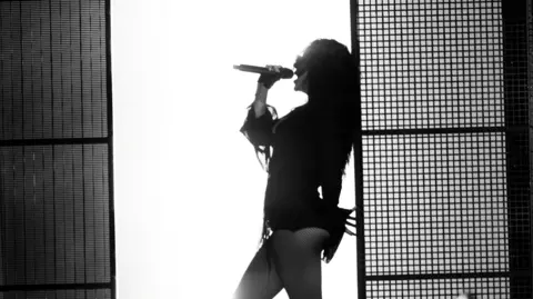 Henry Redcliffe A black and white photo of Charli XCX leaning against a wall as she sings into a microphone. There is a bright white wall behind her which silhouettes her body.