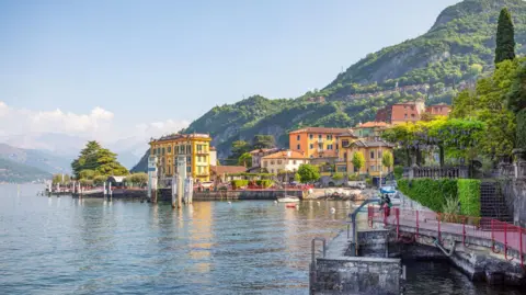 Getty Images Sunny yellow buildings and greenery sit infront of green mountains overlooking Lake Como. 