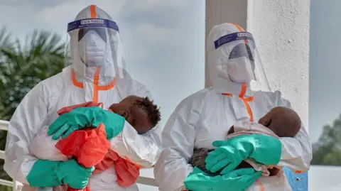 Reuters Two health workers in PPE each hold a young baby. 