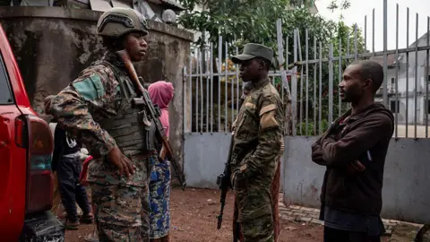 AFP via Getty Images Two men dressed in military uniform and holding rifles stand alongside a member of the public in a brown hoody and trousers. A woman in a pink hoody has her back to the camera in the background