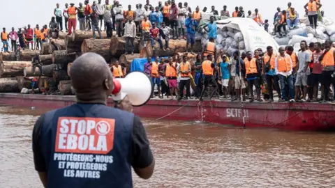 Reuters A man with a megaphone has his back to the camera. The words "Stop Ebola" are visible on the back of his jacket. He is calling to a boat full of people wearing orange life jackets.