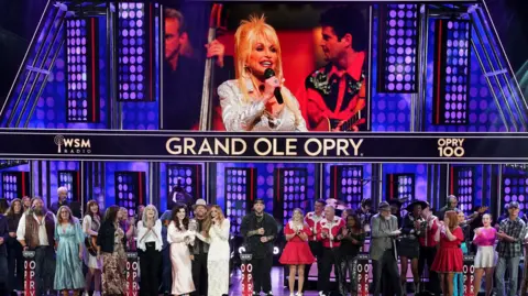 Reuters More than two dozen singers and musicians in a row on stage below a large screen showing a photo of Dolly Parton and the Grand Ole Opry name