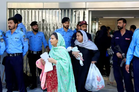 Reuters Two women, one carrying a baby, wearing headscarves leave the hospital ward surrounded by police. 