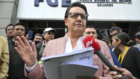 Getty Images Fernando Villavicencio speaks into a microphone while holding papers and gesturing with one hand on the steps of a building surrounded by people. He wears a pink jacket, white shirt and lack-rimmed glasses.
