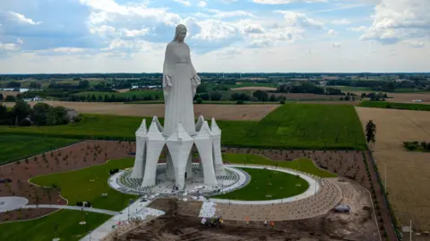 Getty Images A statue of the Virgin Mary rises out of green and yellow fields in Poland on a cloudy sky. 