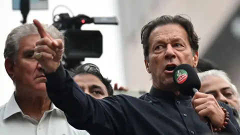 Getty Images Imran Khan wearing a black button-up shirt and speaking into a microphone, points dramatically. Behind him are other men, and a television camera