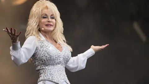 Getty Images Dolly Parton, wearing a white and silver bedazzled outfit, on stage smiling and holding out her arms