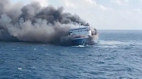 Basarnas A still shot of the KMP Mutiara Sentosa with smoke billowing from behind as the passenger vessel slants to the side in the ocean