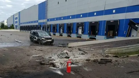 Handout A large building that looks like a warehouse, with multiple entry ports for transport vehicles. The tarmac in front is charred and damaged, and a fire extinguisher has been left at the scene. A car is badly damaged, and the building also appears to have some damage.