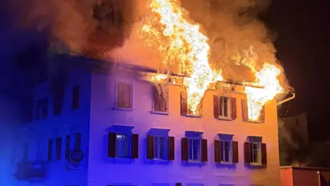 Graubünden cantonal police Large flames come out of the windows in the top floor of a building at night.