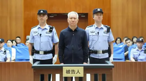 Shenzhen Intermediate People’s Court/WeChat China Evergrande Group founder Hui Ka Yan takes the stand in a court in China. He has a blank look and is standing between two officers, with members of the audience in the background.
