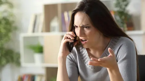 Pheelings Media/ Getty Images Woman with angry expression holds phone up to her ear looking frustrated. She has brown hair and wears a grey knit top.