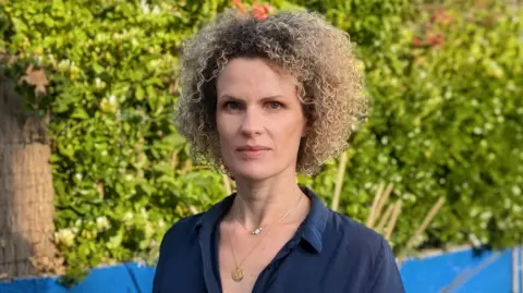 BBC Anaïs de Vos is wearing a dark blue shirt with an open neck and is standing in front of bushes and a low, bright blue wall. She is wearing a gold necklace and has light, curly hair. 
 