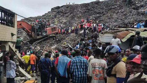 AFP via Getty Images Rescuers use an excavator to comb through a mountain of scattered rubbish at a major waste site in Conakry, Guinea, as local residents gather nearby. Photo: 23 August 2026