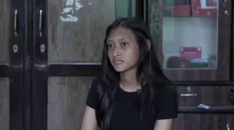 A girl with dark hair sits being interviewed in front of a door, she is in a black t-shirt.