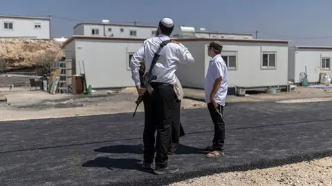 Ilia YEFIMOVICH / AFP via Getty Images Three Israeli settlers stand outside houses after the reopening of a settlement at Ganim in the Israeli-occupied West Bank on 13 August. One is a man armed with a rifle. The image has a blue border with the BBC Verify logo in the the top left corner. 