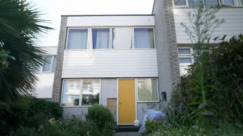 BBC/Kevin Church A terraced house with light grey brick walls, a series of windows on the upper floor, two windows and a yellow door on the ground. There is a garden path and bushes on the side