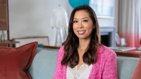 Vivien Wong Vivien Wong wearing a white lace camisole and bright pink wool cardigan sitting on a grey sofa in a plush home