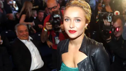 Getty Images Hayden Panettiere surrounded by photographers she has her hair tied back and wearing red lipstick and a greendress with a black leather jacket over the top